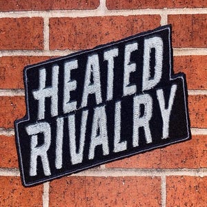 May include: A black patch with the words "HEATED RIVALRY" in white, stylized lettering. The patch has a rectangular shape with a curved bottom edge and is set against a red brick wall.