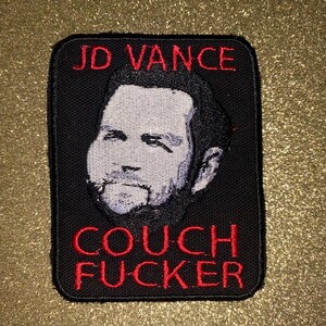 May include: Black rectangular patch with a portrait of a man and the text "JD VANCE" in red at the top. Below the portrait, the text "COUCH FUCKER" is also in red. The patch is on a gold background.