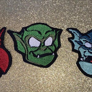 May include: Three embroidered patches featuring cartoon gargoyle faces. The patches are red, green, and blue, each with unique expressions and details. The patches are outlined in black and have white and red accents.