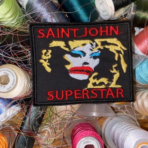 Może przedstawiać: Czarna naszywka haftowana czerwonymi i żółtymi ściegami. Naszywka przedstawia stylizowany obraz osoby z tekstem "SAINT JOHN SUPERSTAR" nad i pod obrazem.