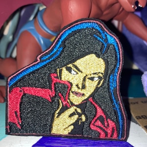 May include: An embroidered patch featuring a stylized illustration of a person with long black hair, a gold-toned face, and red clothing accents. The design is outlined in blue and black thread, creating a detailed portrait with a sharp, defined aesthetic.