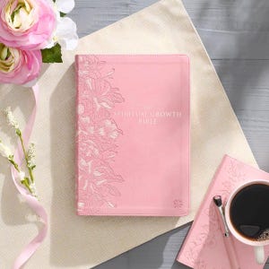May include: A pink Bible with the text "The Spiritual Growth Bible" in white. The cover features a floral design on the left side. A matching pink notebook, pen, and coffee cup are also present, along with pink flowers.