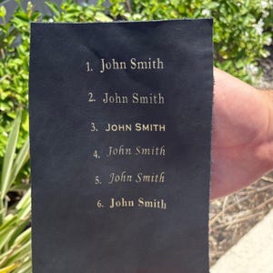May include: A black leather rectangle with gold lettering. The text reads: 1. John Smith, 2. John Smith, 3. JOHN SMITH, 4. John Smith, 5. John Smith, and 6. John Smith. The item is held by a person.