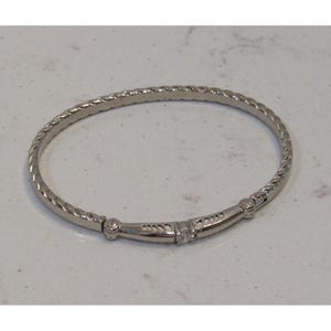 May include: A silver-colored bangle bracelet with a twisted rope design around the band. The bracelet features a decorative center section with small, clear gemstones. The bracelet is designed to be worn on the wrist.