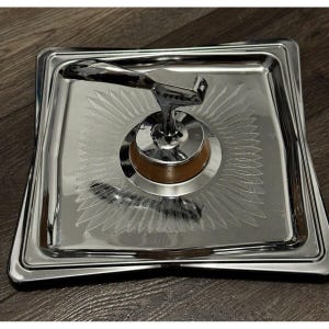 May include: A chrome-plated serving tray with a decorative design. The tray features a central, circular wooden base with a chrome handle. The tray's surface has a sunburst pattern. The tray is square with rounded corners.