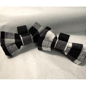 May include: Three black and white checkered bow ties are arranged on a white surface. The bow ties have a classic plaid pattern, with a black band for neck attachment. The fabric appears to be woven.
