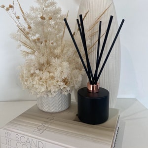 May include: Black diffuser bottle with black reeds and a rose gold cap, sitting on a stack of books with the text "Scandi Coastal Outdoor Living" on the cover. The diffuser is next to a white vase with dried white flowers.