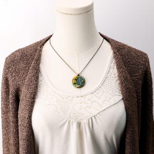 Necklace shown on a white mannequin wearing a cream top and brown sweater. The teal-blue hobbit door pendant with sunflowers, moss ring, and stone doorstep sits at collarbone length on an aged bronze chain.