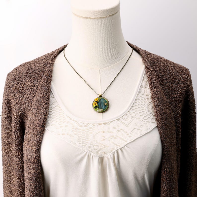 Necklace shown on a white mannequin wearing a cream top and brown sweater. The teal-blue hobbit door pendant with sunflowers, moss ring, and stone doorstep sits at collarbone length on an aged bronze chain.