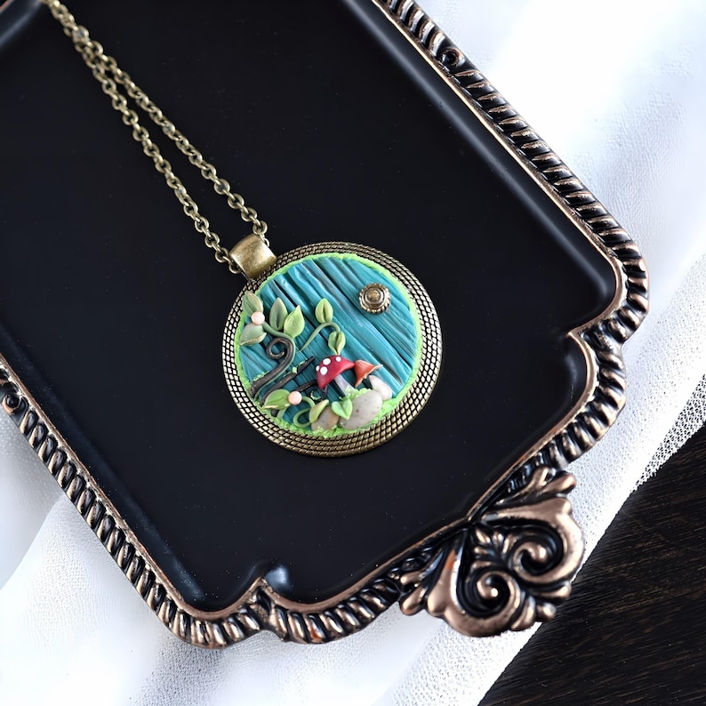 May include: A charming pendant necklace with a miniature forest design. The circular pendant showcases a blue backdrop with a tree, red mushrooms, and a small sun. The pendant is framed in bronze-coloured metal and hangs from a matching chain.