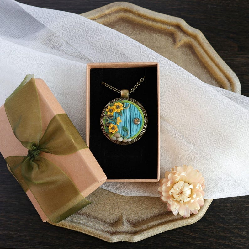 May include: A handcrafted pendant necklace with a round, blue door design, yellow flowers, and a bronze chain. The necklace is in a peach gift box with a green ribbon. A dried flower and a decorative tray are also present. The necklace is a unique piece of jewellery.