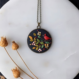 May include: A round pendant necklace with a black background, featuring a red cardinal bird perched on a branch with colorful berries and leaves. The pendant is framed in a bronze-colored metal and hangs from a matching chain.