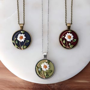 May include: Three daisy pendant necklaces. Each pendant features a white daisy with an orange center, surrounded by green leaves and stems. The pendants have dark blue, burgundy, and olive green backgrounds. The chains are bronze and silver.