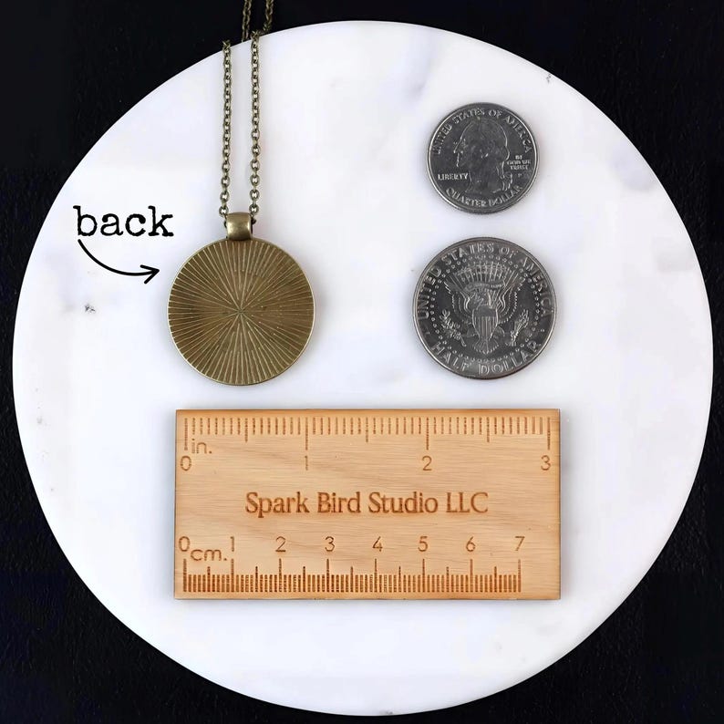 Reverse of pendant displayed with two U.S. coins and a wooden ruler for scale. Shows the textured aged-bronze back of pendant and Spark Bird Studio LLC ruler for approximate 1.25-inch diameter reference. The pendant is larger than a U.S. quarter, it is roughly the size of a U.S. half dollar coin.
