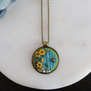 May include: A circular pendant necklace with a blue door design, yellow sunflowers, and green detailing. The pendant hangs from a bronze-toned chain. The door has a textured surface and a small, round doorknob.