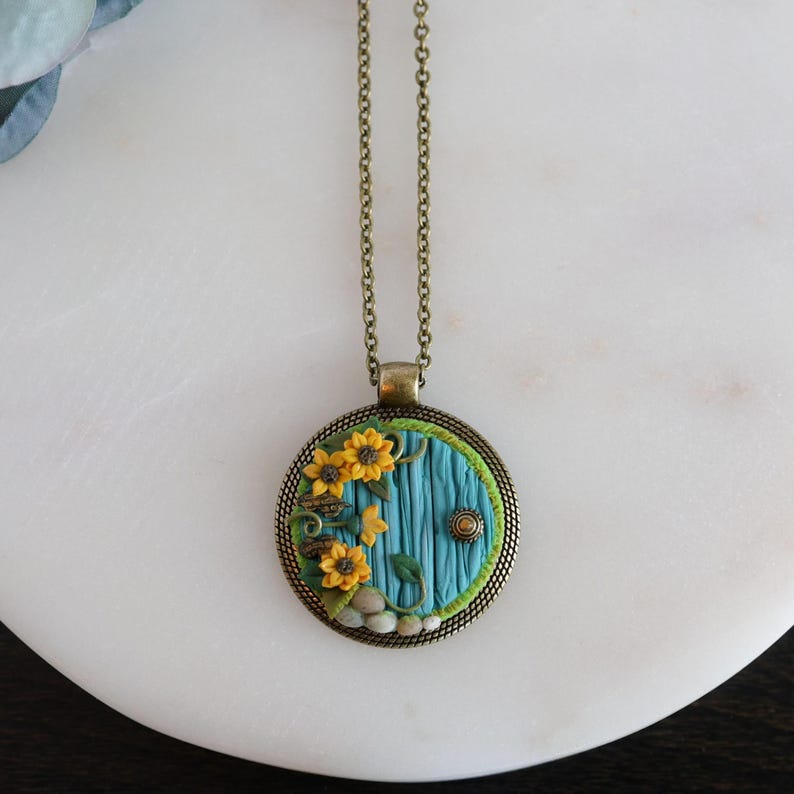 May include: A circular pendant necklace with a blue door design, yellow sunflowers, and green detailing. The pendant hangs from a bronze-toned chain. The door has a textured surface and a small, round doorknob.