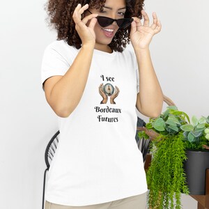 Peut inclure: T-shirt blanc avec un graphique d'une boule de cristal avec des mains la tenant. Le texte sur le t-shirt est "I see Bordeaux Futures".