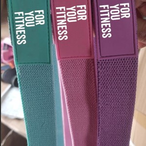 Pode incluir: Tr&ecirc;s bandas de resist&ecirc;ncia de tecido em verde, rosa e roxo com o texto "FOR YOU FITNESS" impresso em cada banda.