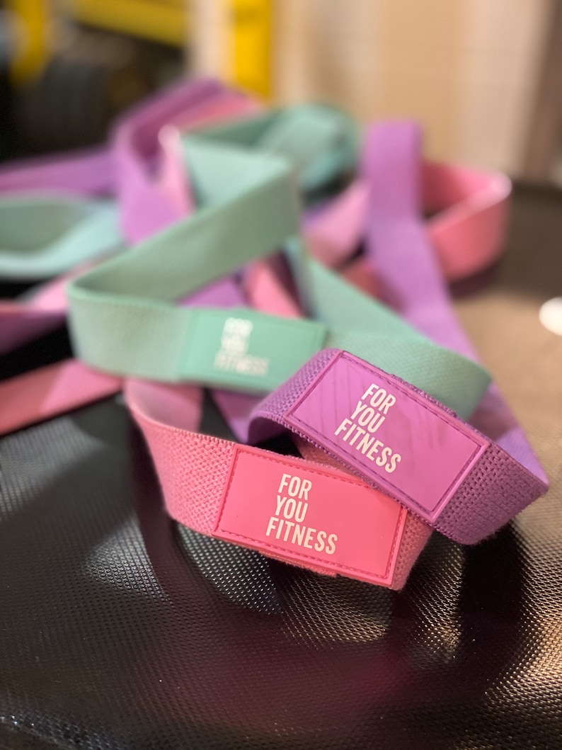 Pode incluir: Um conjunto de faixas de resist&ecirc;ncia em rosa, roxo e verde com o texto "For You Fitness" impresso nas faixas.