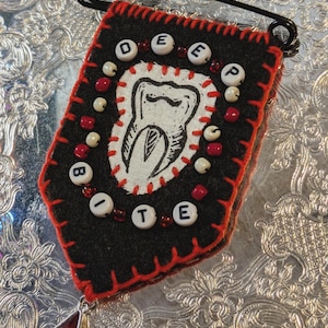 May include: A black felt banner with red stitching and a safety pin. The banner features the words "DEEP BITE" spelled out with white letter beads and a tooth graphic. Red and white beads surround the tooth graphic and the banner's edges. A red teardrop charm hangs from the bottom.