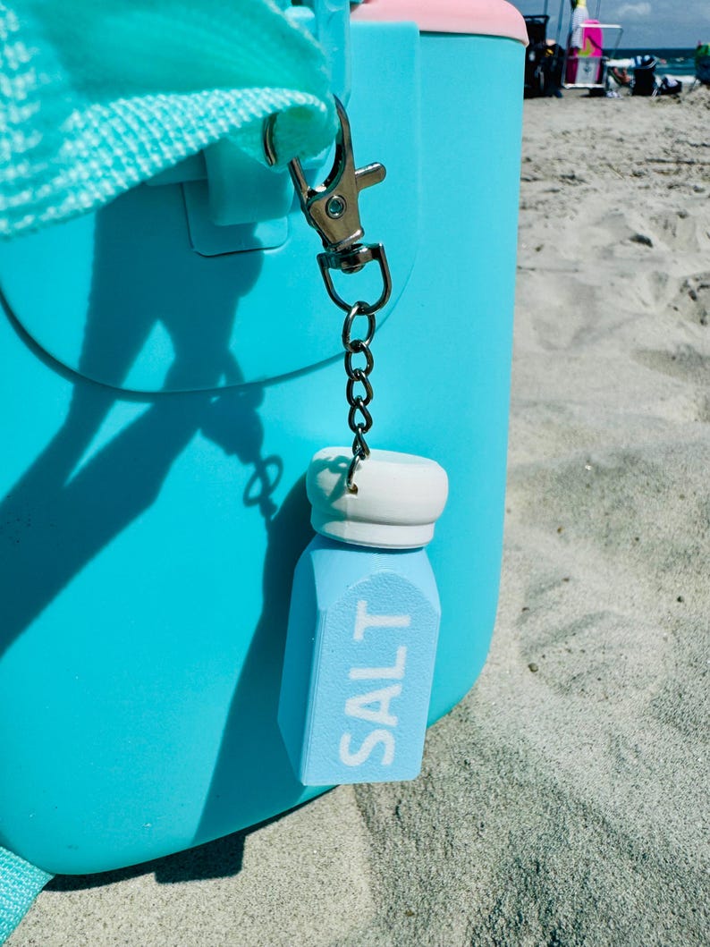 May include: A light blue salt shaker keyring with the word "SALT" in white lettering. The keyring is attached to a silver clasp and chain, hanging from a turquoise cooler. The cooler has a pink top and is on a sandy beach.