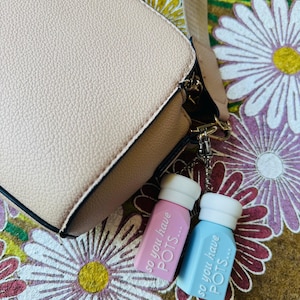 May include: A light pink and light blue keychain with the text "so you have POTS..." hangs from a beige handbag. The handbag has a brown base and a beige strap. The background is a floral pattern.