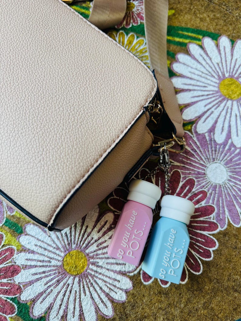 May include: A light pink and light blue keychain with the text "so you have POTS..." hangs from a beige handbag. The handbag has a brown base and a beige strap. The background is a floral pattern.