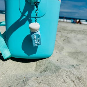 May include: A turquoise bucket with a pink rim and a matching strap sits on the sand at the beach. A small charm with the text "so you have POTS..." hangs from the bucket's handle. The sky is blue with a few clouds.