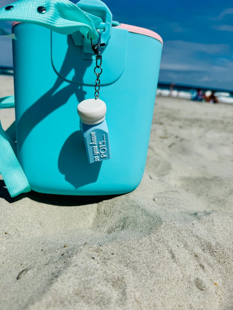 May include: A turquoise bucket with a pink rim and a matching strap sits on the sand at the beach. A small charm with the text "so you have POTS..." hangs from the bucket's handle. The sky is blue with a few clouds.