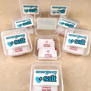Pode incluir: Recipientes de plástico transparente com a etiqueta "emergency salt" e um coração azul e um gráfico de saleiro. Cada recipiente contém pequenos pacotes de "All Natural Sea Salt". Os recipientes estão dispostos sobre uma superfície castanha clara.