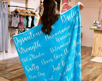 Op de afbeelding: Een felblauwe fleece deken met witte cursieve tekst, waaronder woorden als Dysautonomia, Strength, Perseverance, Warrior, Determined, Not a Burden, Never Give Up, Salty, Flare Day, It's okay to rest, Horizontal en Exhaustion.