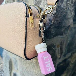 May include: A pink salt shaker charm with the word "SALT" in white lettering, attached to a light tan handbag. The bag has gold-coloured zips and hardware. The charm has a white top and is on a silver chain.