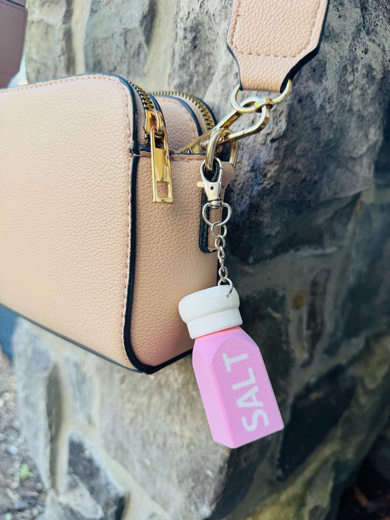 May include: A pink salt shaker charm with the word "SALT" in white lettering, attached to a light tan handbag. The bag has gold-coloured zips and hardware. The charm has a white top and is on a silver chain.
