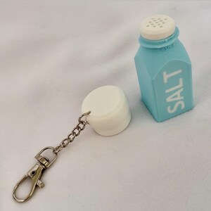 May include: A novelty salt shaker keychain. The light blue, carton-shaped shaker has a white top and the word "SALT" printed on the side. A white, cylindrical cap is attached to the shaker with a chain and a silver-coloured clasp.