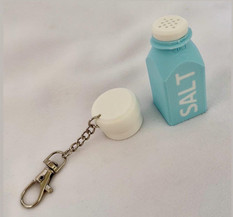 May include: A novelty salt shaker keychain. The light blue, carton-shaped shaker has a white top and the word "SALT" printed on the side. A white, cylindrical cap is attached to the shaker with a chain and a silver-coloured clasp.