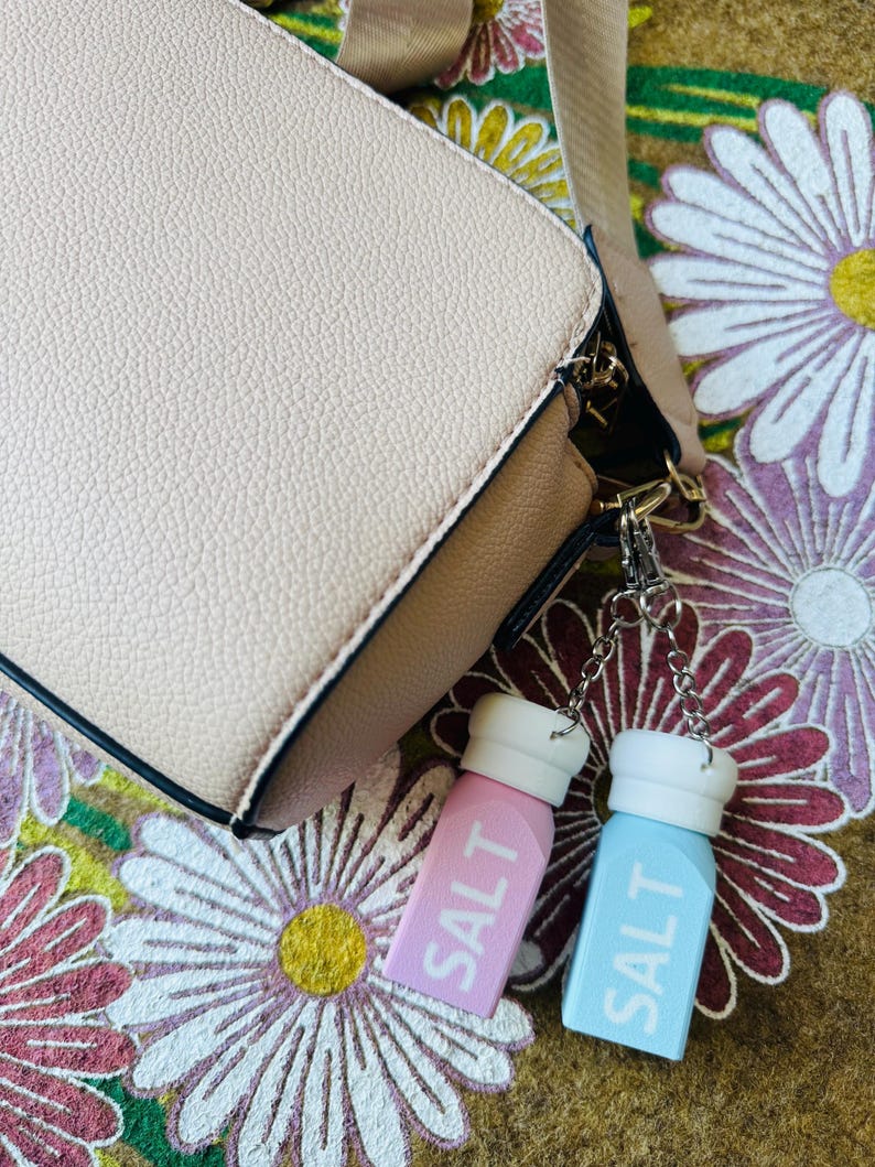 May include: A pale pink crossbody bag with a beige strap and two salt shaker keyrings. One keyring is light pink with white text that reads "SALT", and the other is light blue with white text that reads "SALT".