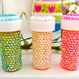 May include: Three decorative, bedazzled bottles in shades of green, peach, and pink. Each bottle is covered in iridescent rhinestones and topped with a white lid. The bottles are arranged on a white surface with floral accents.