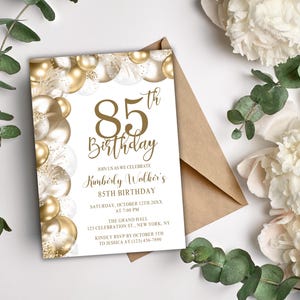 Puede incluir: Una invitación para un 85 cumpleaños. La tarjeta presenta globos dorados y el texto "85 cumpleaños", así como los detalles del evento. La invitación va acompañada de un sobre marrón y flores blancas.