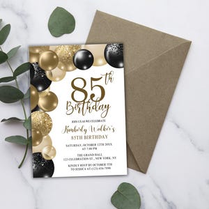 Puede incluir: Una invitación de cumpleaños número 85 con globos dorados, negros y blancos. La invitación incluye el texto "85th Birthday" y los detalles del evento. Un sobre marrón está al lado de la invitación.