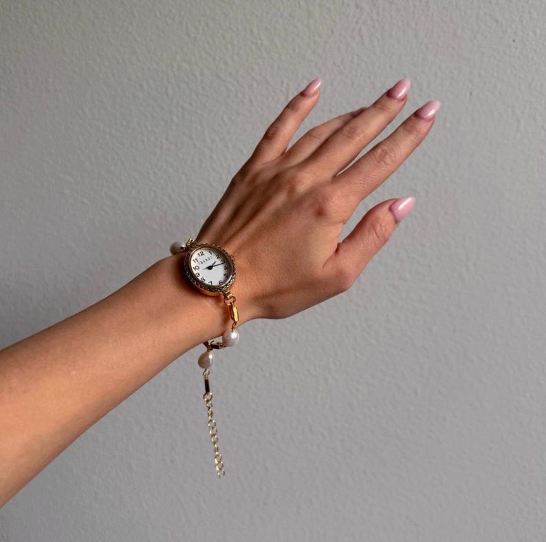 K&ouml;nnte beinhalten: Eine goldfarbene Uhr mit wei&szlig;em Zifferblatt und schwarzen Ziffern ist an einem Perlenarmband befestigt. Die Uhr hat eine dekorative L&uuml;nette und wird am Handgelenk getragen. Der Hintergrund ist ein neutrales Grau.