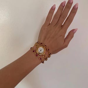 K&ouml;nnte beinhalten: Goldfarbene Armbanduhr mit wei&szlig;em Zifferblatt und schwarzen Zeigern. Die Uhr ist von einem doppelreihigen Armband mit roten Edelsteinen umgeben. Die Uhr befindet sich an einem Handgelenk.