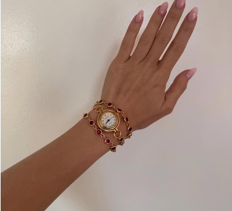 K&ouml;nnte beinhalten: Goldfarbene Armbanduhr mit wei&szlig;em Zifferblatt und schwarzen Zeigern. Die Uhr ist von einem doppelreihigen Armband mit roten Edelsteinen umgeben. Die Uhr befindet sich an einem Handgelenk.