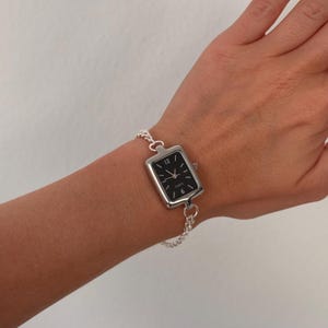 May include: A silver-toned rectangular watch with a black face and white hour markers. The watch is attached to a delicate chain bracelet. The watch face displays the numbers 12, 3, 6, and 9.