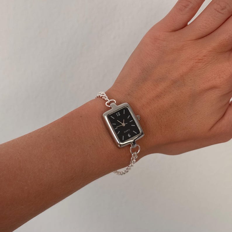 May include: A silver-toned rectangular watch with a black face and white hour markers. The watch is attached to a delicate chain bracelet. The watch face displays the numbers 12, 3, 6, and 9.
