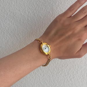 K&ouml;nnte beinhalten: Eine goldfarbene Armbanduhr mit wei&szlig;em Zifferblatt und schwarzen Ziffern. Die Uhr hat ein ovales Zifferblatt und ist an einem goldenen Kettenarmband befestigt. Die Uhr befindet sich an einem Handgelenk.