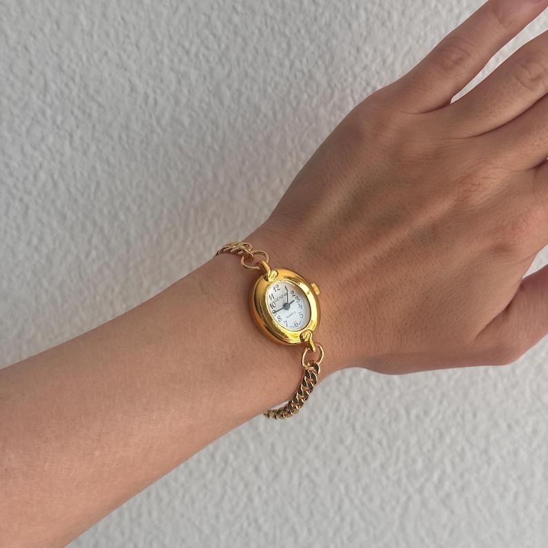 K&ouml;nnte beinhalten: Eine goldfarbene Armbanduhr mit wei&szlig;em Zifferblatt und schwarzen Ziffern. Die Uhr hat ein ovales Zifferblatt und ist an einem goldenen Kettenarmband befestigt. Die Uhr befindet sich an einem Handgelenk.