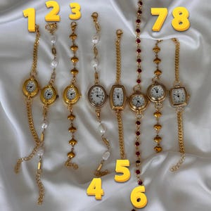 K&ouml;nnte beinhalten: Acht goldfarbene Armbanduhren mit unterschiedlichen Designs. Einige haben wei&szlig;e Zifferbl&auml;tter mit schwarzen Ziffern, andere dekorative Ketten, Perlen und herzf&ouml;rmige Akzente. Die Uhren sind auf einem wei&szlig;en, seidigen Hintergrund pr&auml;sentiert.