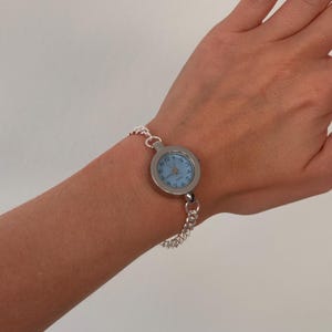 May include: A silver-toned watch with a light blue face and silver chain bracelet. The watch face has black numerals and hands. The watch is a classic, elegant accessory.