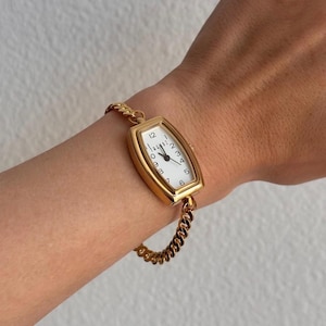K&ouml;nnte beinhalten: Eine goldfarbene Armbanduhr mit rechteckigem Zifferblatt und wei&szlig;em Zifferblatt. Die Uhr hat schwarze Ziffern und Zeiger und ist an einem goldenen Kettenarmband befestigt. Die Uhr wird am Handgelenk getragen.