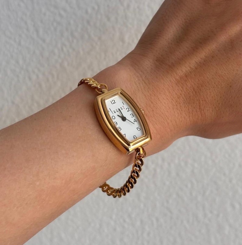 K&ouml;nnte beinhalten: Eine goldfarbene Armbanduhr mit rechteckigem Zifferblatt und wei&szlig;em Zifferblatt. Die Uhr hat schwarze Ziffern und Zeiger und ist an einem goldenen Kettenarmband befestigt. Die Uhr wird am Handgelenk getragen.
